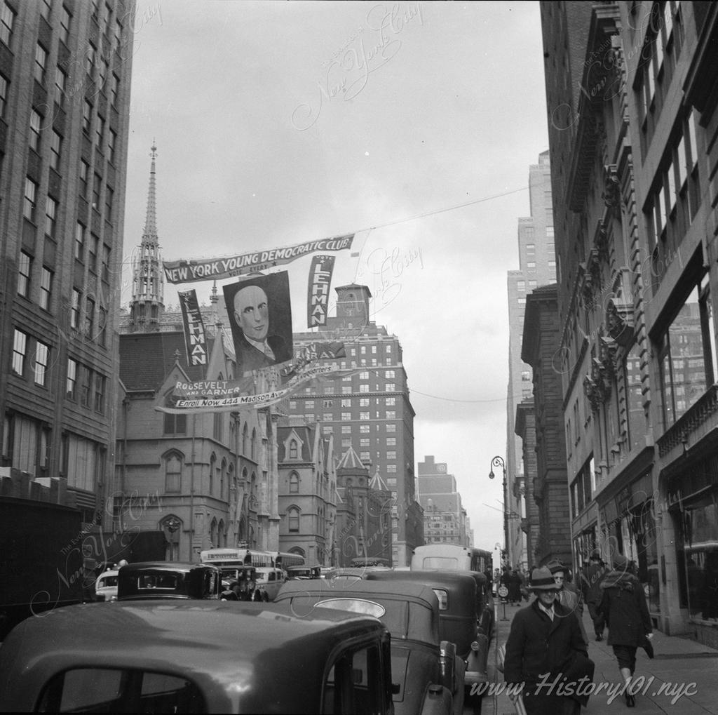 Photograph looking up Madison Avenue, north of 49th Street, full of cars and pedestrians with a political banner for the New York Young Democratic Club hanging overhead.