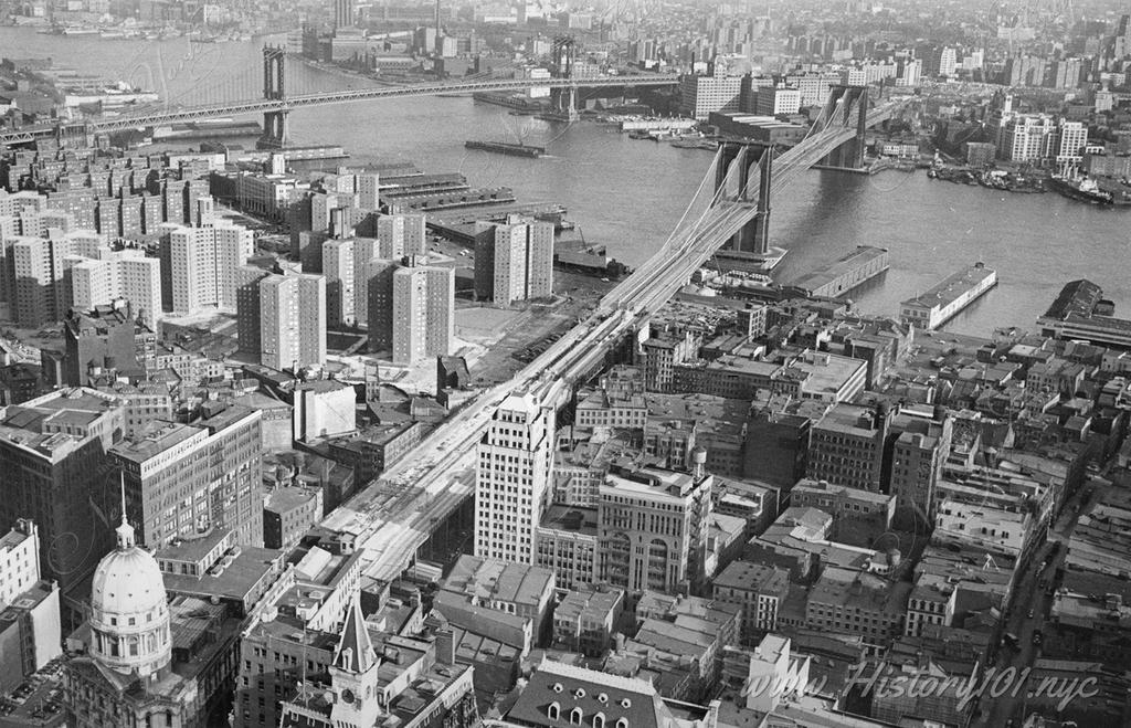 Photograph shows an aerial view of Manhattan including the East River featuring the Manhattan and Brooklyn bridges.