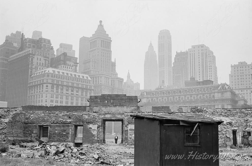 Photograph of a man standing in the doorway of a building in ruins against a backdrop of skyscrapers.